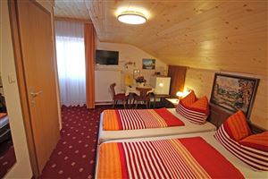 A cozy bedroom with two beds and colorful pillows. There is a small table, a TV, and plenty of natural light through the window.