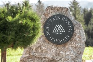Ein Schild aus Stein mit dem Namen "Mountain Resort Seiwald" und einem einfachen Logo. Die Umgebung zeigt grüne Bäume und eine natürliche Landschaft.