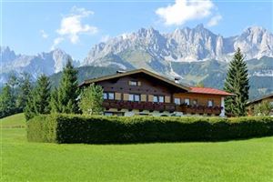 A traditional house surrounded by a green meadow and trees. In the background, majestic mountains rise under a blue sky.