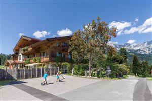 A beautiful wooden house surrounded by trees and mountains. Children ride their scooters on a quiet path.