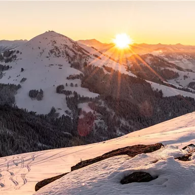 A stunning winter landscape with snow-covered mountains. The sun rises on the horizon, bathing the scene in warm light.