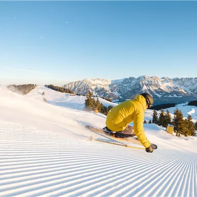 Ein Skifahrer trägt eine gelbe Jacke und fährt die perfekt präparierte Piste hinunter. Im Hintergrund sind schneebedeckte Berge und ein klarer blauer Himmel zu sehen.