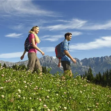 A couple is hiking in a green meadow full of flowers with mountains in the background. The sky is clear and blue with a few clouds.