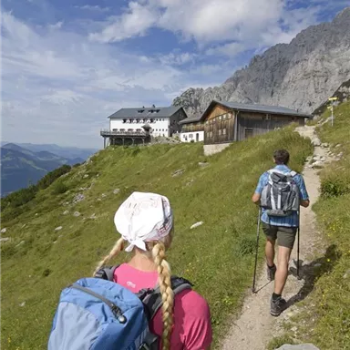 Two hikers on a path in the mountains, with a view of a cabin in the background. The landscape is green and the clouds are partially visible in the sky.