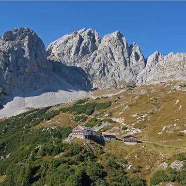 Eine beeindruckende Berglandschaft mit hohen Gipfeln und grünem Gras. Im Vordergrund befindet sich eine Berghütte.