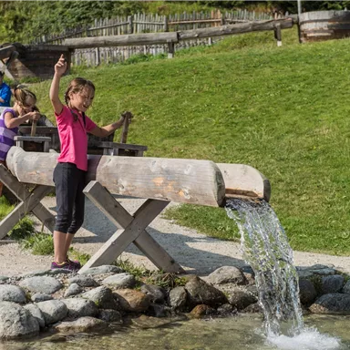 Children are playing at a water play area and experimenting with water. In the background, green meadows and wooden structures can be seen.