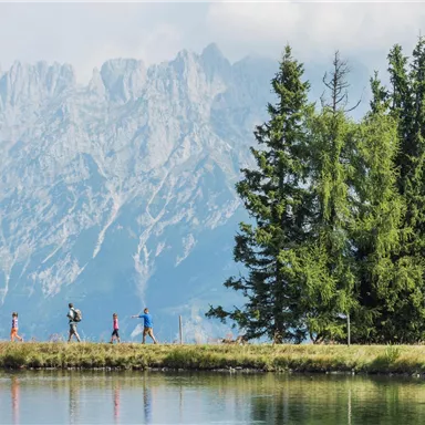 A group of hikers is walking along the shore of a lake. In the background, majestic mountain peaks and green trees are visible.