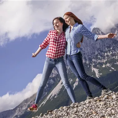 Zwei fröhliche junge Frauen stehen auf einem Kiesfeld und lachen. Im Hintergrund sind majestätische Berge und ein blauer Himmel mit einigen Wolken zu sehen.