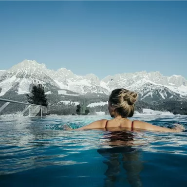 Ein Frau schwimmt in einem Außenpool mit Blick auf schneebedeckte Berge. Der Himmel ist klar und blau.