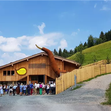 Ein Holzgebäude mit einem eindrucksvollen Hornanbau, umgeben von grünen Wiesen. Eine große Gruppe von Menschen steht vor dem Gebäude und genießt den Tag.