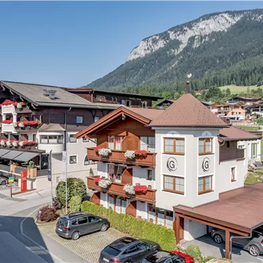A picturesque Alpine landscape with several buildings in the foreground. The surroundings are green and mountainous, ideal for vacationers.