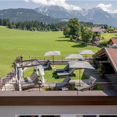 A terrace with loungers and sun umbrellas, surrounded by green meadows and mountains. In the background, a clear sky can be seen.