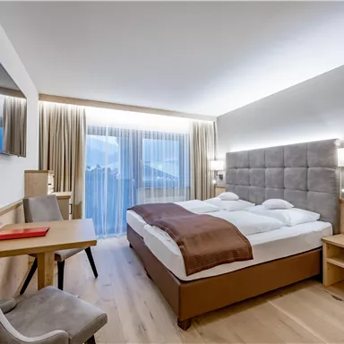 A modern hotel room with a double bed and a desk. The large windows let in a lot of light into the room.
