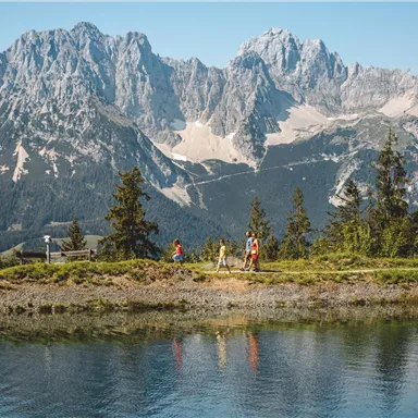Eine malerische Berglandschaft mit hohen Gipfeln und klarem Wasser im Vordergrund. Ein paar Menschen genießen die Natur am Ufer.