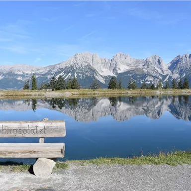 Eine Holzbank am ruhigen See mit Bergen im Hintergrund. Das Wasser spiegelt die umliegende Landschaft und den klaren Himmel wider.