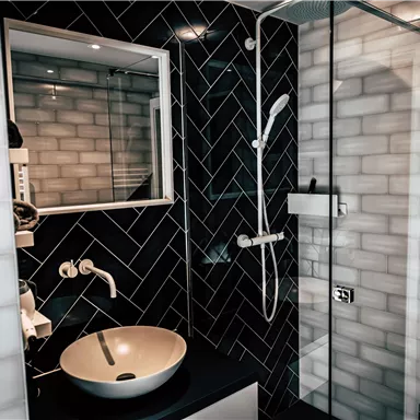 A modern bathroom with black tiles and a large shower cabin. In the center, there is an elegant sink.