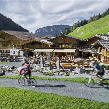 Zwei Radfahrer fahren auf einem Weg vorbei an einem traditionellen Berggasthaus. Im Hintergrund sind grüne Hügel und Berge zu sehen.