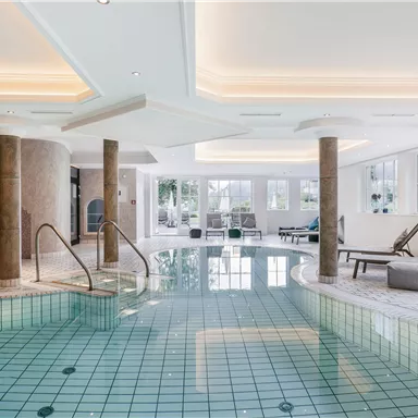 A luxurious indoor pool with a smooth, green tiled floor. The environment is bright and modern, with large windows and comfortable loungers.