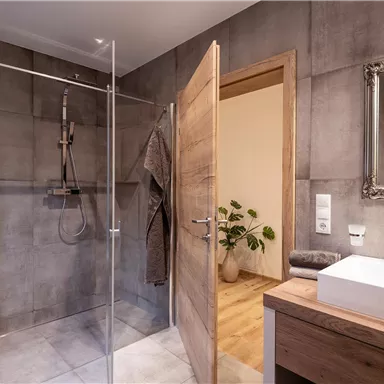 Ein modernes Badezimmer mit einer Glasdusche und einem Holzwaschbecken. Die Wände sind in betongrau gehalten, und im Hintergrund steht eine Pflanze.