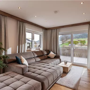 Ein modernes Wohnzimmer mit einem beigen Sofa und großen Fenstern. Die Aussicht zeigt eine grüne Landschaft und Berge im Hintergrund.