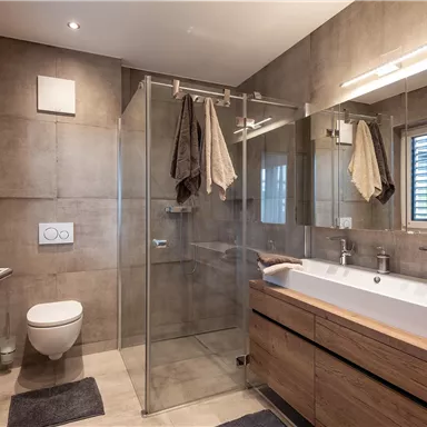 A modern bathroom with a glass shower cabin and wooden shelves. The walls are in neutral colors, and there are large windows for natural light.