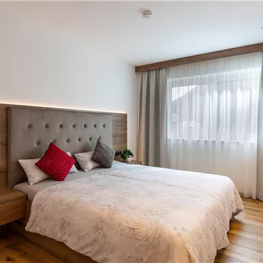 A modern bedroom with a large double bed and an elegant headboard. The room is bright and cheerful, with wooden flooring and curtains made of light fabric.