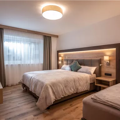 A modern hotel room with two beds and a wooden floor. The room has large windows with curtains and a warm, inviting lighting.