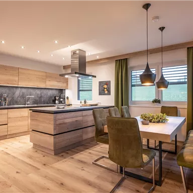A modern kitchen with wooden shelves and a big dining table. The open atmosphere is emphasized by the elegant lamps and the green curtains.