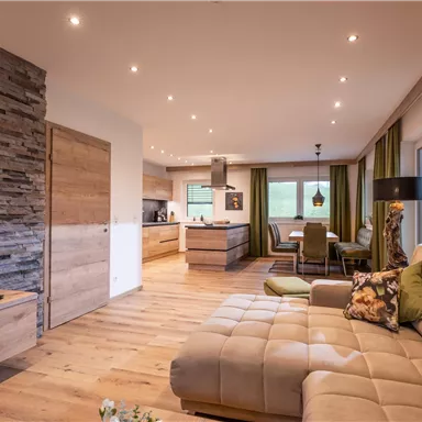 A modern living room with a comfortable sofa and wood-paneled walls. The open kitchen is bright and inviting, with large windows and plenty of daylight.