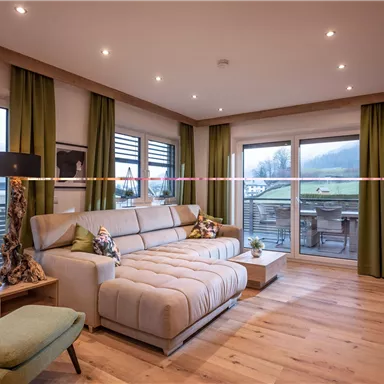Eine moderne Wohnstube mit hellem Holzfußboden, gemütlichem Sofa und großen Fenstern. Die grünen Vorhänge und der Blick nach draußen schaffen eine einladende Atmosphäre.