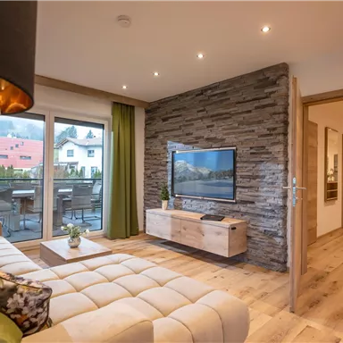 Ein modernes Wohnzimmer mit einem gemütlichen Sofa und einem Fernseher. Große Fenster bieten Blick auf einen Balkon und viel Tageslicht.