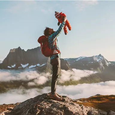 Ein Mensch hebt ein Kind auf einem Berggipfel in die Luft. Im Hintergrund sind beeindruckende Berge und Wolken zu sehen.