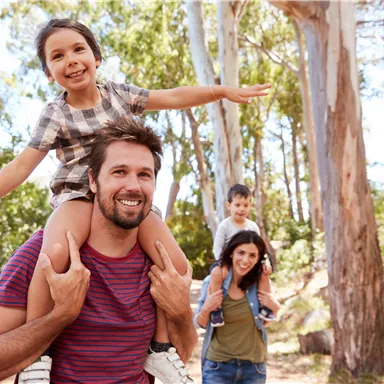 Eine Familie genießt einen Ausflug in der Natur. Ein Vater trägt seine Tochter auf den Schultern, während die Mutter und der Sohn im Hintergrund stehen.