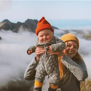 Eine lächelnde Mutter hält ein Baby in den Armen, beide tragen bunte Mützen. Im Hintergrund sind Berge und eine Wolkendecke zu sehen.