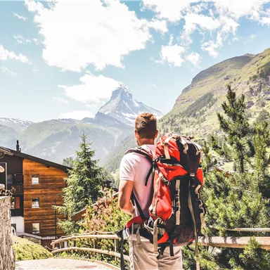 Ein Wanderer mit einem Rucksack steht vor einer beeindruckenden Berglandschaft. Im Hintergrund ist der Matterhorn sichtbar, umgeben von grünen Hügeln und klarer Blauen Himmel.