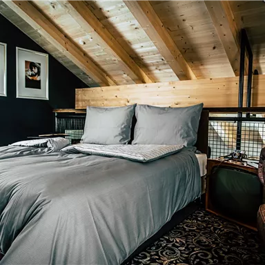Ein stilvolles Schlafzimmer mit einem großen Bett und einem modernen Braun-Lederstuhl. Die Holzbalkendecke und die Bilder an der Wand verleihen dem Raum Wärme und Gemütlichkeit.