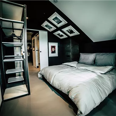 Ein modernes Schlafzimmer mit einem großen Bett und minimalistischer Einrichtung. Es gibt eine Leiter und mehrere Bilder an der Wand.