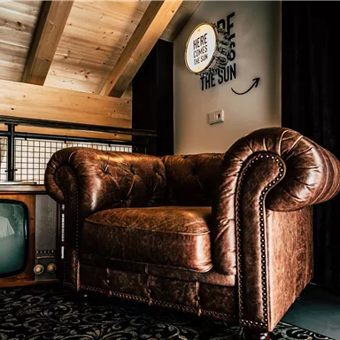Ein gemütlicher, brauner Ledersessel steht in einem modernen Raum. Im Hintergrund befindet sich ein retro Fernseher und eine schöne Holzdecke.
