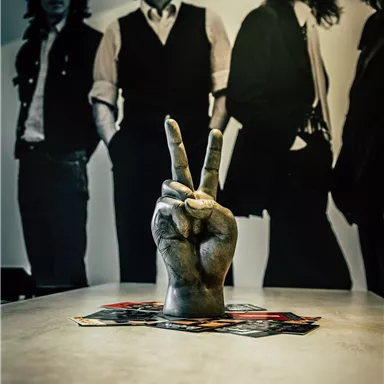 Eine Hand mit dem Peace-Zeichen steht auf einem Tisch. Im Hintergrund sind Silhouetten von vier Männern zu sehen.