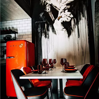 A modern dining room with a white table and black chairs. An impressive mural hangs on the wall, while a red refrigerator stands in the background.
