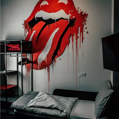 A creative mural of a red lips design with a sticking-out tongue. In the room stands a black bed with pillows and a shelving structure.