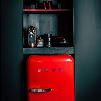A red refrigerator from SMEG stands in a dark kitchen environment. Next to it, some kitchen appliances and dishes can be seen on a shelf.