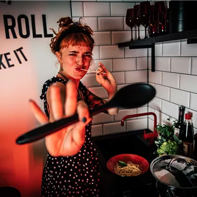 Eine fröhliche Frau kocht in einer modernen Küche. Sie hält einen Löffel in der Hand und scheint Spaß beim Kochen zu haben.