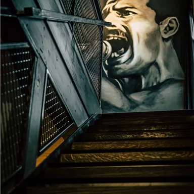 Eine Treppe mit dunklem Holz und einem metallischen Geländer. An der Wand hängt ein beeindruckendes Wandgemälde eines schreienden Mannes.