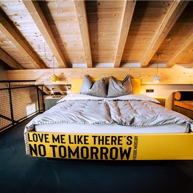 Ein modernes Schlafzimmer mit einem großen Bett und Holzdecken. Die Bettkante hat einen auffälligen Schriftzug: "LOVE ME LIKE THERE'S NO TOMORROW".