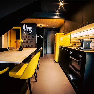 A modern kitchen with yellow chairs and black furniture. The walls are stylishly designed, featuring a striking refrigerator and warm lighting.
