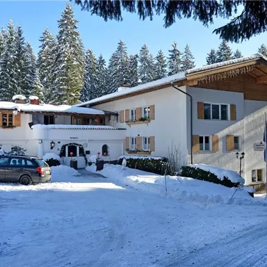 Ein gemütliches Gebäude im Winter, umgeben von schneebedeckten Bäumen. Der Himmel ist klar und blau, und ein Auto steht vor dem Haus.