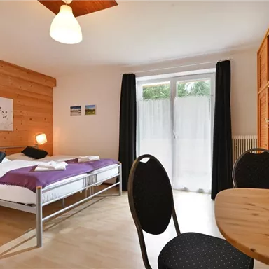 Ein gemütliches Zimmer mit einem Doppelbett, Holzverkleidung und großen Fenstern. Ein runder Tisch mit Stühlen und einer Vase voller Blumen unterstreicht die einladende Atmosphäre.