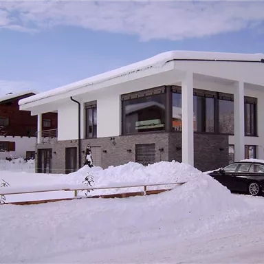 Ein modernes Haus inmitten einer verschneiten Landschaft. Die Umgebung ist ruhig mit schneebedeckten Bäumen und einer klaren Winterhimmel.