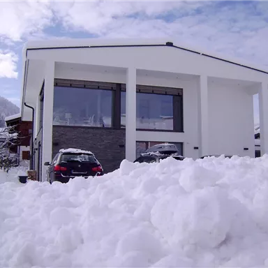 Ein modernes Haus in einer verschneiten Landschaft. Der Schnee türmt sich vor dem Gebäude und der Himmel ist teilweise bewölkt.